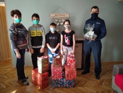Zdjęcie grupowe dzieci, kobiety oraz umundurowanego policjanta. Przed nimi paczki z prezentami