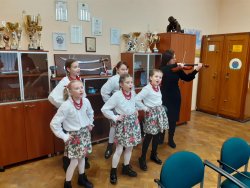Dzieci śpiewające w strojach ludowych obok kobieta gra na skrzypcach
