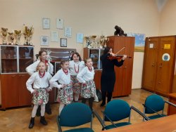 Dzieci śpiewające w strojach ludowych obok kobieta gra na skrzypcach