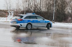 Fotografia przedstawia radiowóz policyjny marki Kia Ceed o nr rej HPGC-143 i numerze taktycznym G484 podczas wykonywania ćwiczenia na płycie poślizgowej. Widoczny od tyłu samochód znajduje się w poślizgu nadsterownym. Na podłożu widać cienką taflę wody.