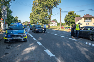 Policjanci zatrzymują samochody do kontroli, obok stoi kobieta z pakietami promocyjnymi.