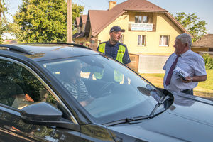 Policjant prowadzi rozmowę z mężczyzną trzymającym ulotki informacyjne, podczas gdy w zatrzymanym do kontroli samochodzie siedzi kobieta.