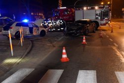 ogólne miejsce kolizji. W tle wóz bojowy straży pożarnej stojący bokiem, przed nim grupa strażaków. W centralnej części zniszczony szary samochód (CitroenC5). Zniszczenia w przedniej części, po lewej stronie biały samochód osobowy audi stojący tyłem w słupkach oddzielających jezdnię od chodnika. Na pierwszym planie wydzielone miejsce kolizji pachołkami drogowymi. Zdjęcie zrobione nocą.