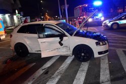 Zniszczenia samochodu audi z prawej strony. Pojazd ma podniesioną pokrywę silnika w tle radiowóz i pojazd straży pożarnej na sygnałach uprzywilejowania. Zdjęcie zrobione nocą.