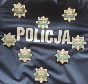 Na granatowym tle z kurtki policyjnej ułożone w kształt serca policyjne odznaki z numerami służbowymi. Wewnątrz napis Policja