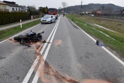 Miejsce wypadku motocyklisty. Na środku drogi rozbity motocykl. Ślady rozsypanego detergentu oczyszczającego drogę z paliwa i oleju. W tle służby ratunkowe.