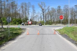 Zdjęcie z drogi podporządkowanej, widoczny znak stop, na drodze ustawione pachołki zamykające ją, w tle widoczny ciemny samochód osobowy, wóz strażacki i stojących obok strażaków