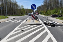 Miejsce wypadku, po prawej stronie rozbity motocykl,  dookoła porozrzucane plastikowe elementy motoru, po prawej stronie na drodze podporządkowanej ciemny samochód osobowy, z tyłu widoczna karetka pogotowia oraz wóz strażacki