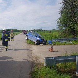 Ogólne miejsce wypadku. Po lewej stronie strażacy, po prawej niebieski samochód osobowy z rozbitą pokrywą silnika, samochód wbity przednią prawą stroną w drogę poprzeczną (mostek)