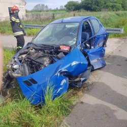 Widok przedniej lewej strony niebieskiego samochodu osobowego, uszkodzona pokrywa silnika, zgniecione i uszkodzone drzwi od kierowcy, wybita szyba w drzwiach kierowcy, po prawej stronie samochodu stoi strażak