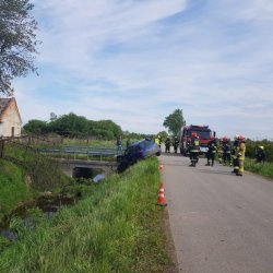 Widok miejsca wypadku z tyłu, po prawej stronie wóz strażacki i stojący dookoła strażacy, po lewej stronie mostek i przechylony w kierunku rowu melioracyjnego niebieski samochód