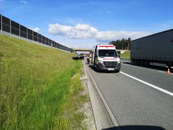 Miejsce wypadku. Na pierwszym planie karetka pogotowia. W tle czarny samochód leżący w rowie na dachu.