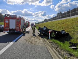 Strażacy sprawdzający wnętrze czarnego samochodu leżącego na dachu w rowie wzdłuż drogi . Po prawej stronie na pasie ruchu ustawiony wóz bojowy straży pożarnej.