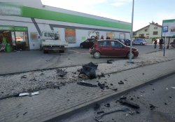 Ogólny widok miejsca wypadku. Widoczne rozrzucone elementy plastikowe od samochodów na chodniku i przestrzeni oddzielającej chodni od parkingu sklepowego. Po prawej uszkodzony bordowy samochód osobowy renault. W centralnej części zdjęcia samochód ciężarowy stojący skośnie do ściany sklepu. Po prawej stronie widocznie ludzie obserwujący zaparkowane samochody przed sklepem a także strażacy zabezpieczający miejsce wypadku. Widoczny również na końcu parkingu po prawej stronie radiowóz policyjny. Elementy tablic rejestracyjnych oraz reklam na sklepie zamazane.