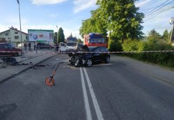 Ogólne miejsce wypadku. Na pierwszym planie widoczny wózek do mierzenia odległości . Miejsce wypadku odseparowane biało-czerwoną taśmą, za którą widoczny jest zniszczony czarny seat. Za nim po prawej stronie wóz bojowy straży pożarnej, po lewej na pasie drogi biały dostawczy Opel. Z lewej strony fotografii chodni, a następnie parking na którym ustawione są samochody osobowe, skośnie. Widoczny słupek przystanku autobusowego.