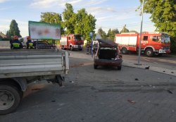 Na zdjęciu po lewej stronie widoczny narożnik samochodu ciężarowego. W centralnym miejscu tył samochodu Renault Clio z podniesionym bagażnikiem i widocznymi uszkodzeniami tylnej części pojazdu. Dalej wozy bojowe straży pożarnej. Policjant rozmawiający z grupa osób.