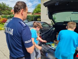 Policjant stoi przy aucie z otwartym bagażnikiem, trójka chłopców wyciąga z bagażnika prezenty