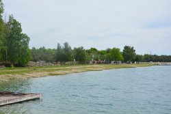 Widoczny zbiornik wodny, a zanim kawałek plaży i droga gruntowa. W tle widoczne drewniane budynki i ogrodzenie z siatki. Za siatka widoczne usypane pagórki ziemi.