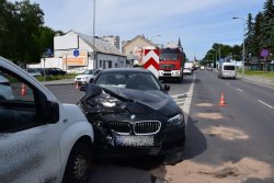 Uszkodzona prawa strona czarnego BMW, rozbita pokrywa silnika, w tle widoczny wóz strażacki i samochody