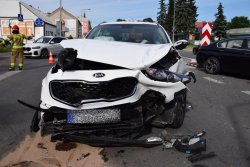 Uszkodzona pokrywa silnika białego samochodu kia, oderwany zderzak oraz rozbite lampy, plastikowe elementy leżą na ziemi
