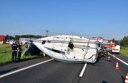 Łódź żaglowa leżąca na lewym boku tarasująca drogę. Po lewej stronie dwóch policjantów mundurowych.