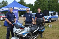 Trzech umundurowanych policjantów stoi przy motorze policyjnym, w tle samochód policyjny oraz wóz strażacki