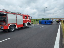 Auto straży pożarnej i przed nim niebieski  leżący na lewy boku samochód. Widoczny spód wywróconego pojazd.