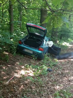 Zielona mazda stojąca na poboczu z otwartym bagażnikiem. Pojazd uderzył w drzewo ma otwarte drzwi przednie prawe