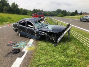 Zniszczone granatowe bmw stojące w poprzek drogi na linii oddzielającej pas ruchu od pobocza, z tyłu widoczne wozy bojowe straży pożarnej