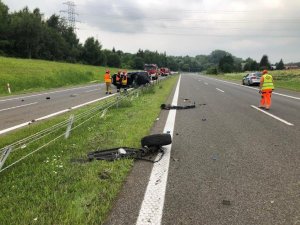 Ogólne miejsce wypadku, na drodze porozrzucane elementy samochodu, z tyłu widoczny ciemny samochód leżący na boku, obok strażacy