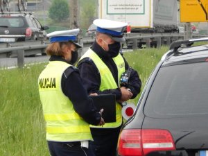 Policjant i policjantka wydziału ruchu drogowego w maseczkach podczas kontroli pojazdu. Na łokciu policjanta oparty laser do mierzenia prędkości