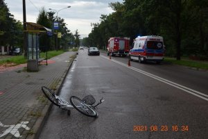 ogólne miejsce wypadku, z przodu widoczny rower leżący na drodze, z tyłu czarny samochód osobowy, po prawej stronie karetka pogotowia i wóz strażacki