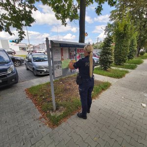 dzielnicowy z Tarnowa zawiesza ulotkę na tablicy ogłoszeń, w tle ulica i supermarket