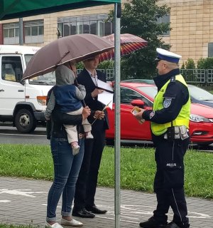 Policjant ruchu drogowego rozmawia z uczniem oraz inna osoba niosącą na ręku małe dziecko. Obie postaci pod parasolami. Policjant wręcza im ulotkę oraz taśmę odblaskową. W tle stojące samochody.
