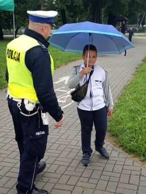 Policjant ruchu drogowego rozmawia z uczniem stojącym na chodniku pod parasolką