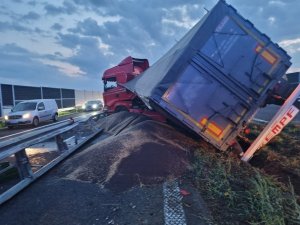 Naczepa samochodu ciężarowego przekręcona na lewą stronę na pas zieleni, sypka zawartość naczepy wysypana na drogę, zniszczone bariery energochłonne