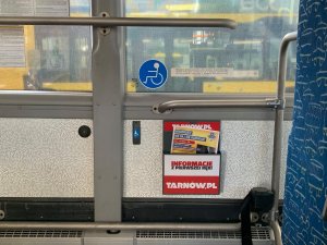 Zdjęcie wnętrza autobusu z miejscami przeznaczonymi do stania. Pod szyba umieszczona skrzynka reklamowa, a w niej plakat w kolorze żółto-niebiesko-czerwonym z dużym napisem „seniorze nie daj się oszukać” z grafiką oraz treścią.