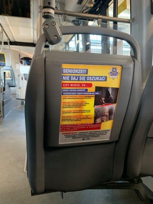 Zdjęcie przedstawia tył fotela autobusowego na którym widniej plakat w kolorze żółto-niebiesko-czerwonym z dużym napisem „seniorze nie daj się oszukać” z grafiką oraz treścią.