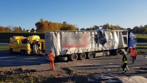 Samochód ciężarowy z rozerwaną plandeką, dookoła mężczyźni zbierający porozrzucane drewniane elementy, za samochodem ciężarowym dwa żółte samochody pomocy drogowej