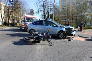 Ogólne miejsce wypadku, na ziemi leży motocykl, obok jasny samochód z uszkodzoną pokrywą silnika, dookoła porozrzucane plastikowe elementy samochodu