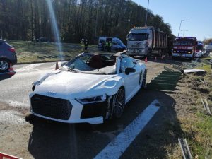 Uszkodzone białe audi, po prawej stronie uszkodzone liny i słupki oddzielające pasy ruchu, z tyłu widoczne wozy strażackie, samochód policyjny oraz auto ciężarowe