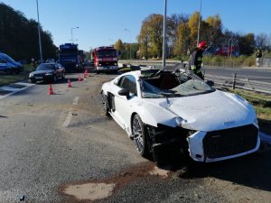 Uszkodzone białe audi, obok strażak, przed samochodem rozsypany piasek neutralizujący oleje i paliwo, z tyłu widoczne samochody oraz wóz strażacki