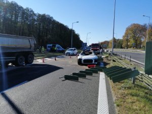 Ogólne miejsce wypadku, uszkodzone i wywrócone liny oraz metalowe słupki oddzielające pasy ruchu, z tyłu widoczny samochód policyjny oraz wóz strażacki