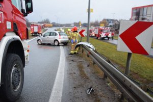 miejsce wypadku, srebrny samochód stojący w poprzek pasa ruchu, obok samochodu stoją strażacy, widoczne wozy bojowe straży pożarnej