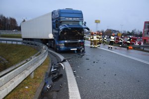 Samochód ciężarowy z uszkodzonym przednim pasem, na drodze porozrzucane plastikowe elementy, po prawej stronie strażacy