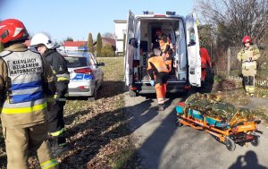 Karetka pogotowia z ratownikami medycznymi w środku, przed karetką nosze z osobą okrytą folią termiczną, wokół stoją strażacy