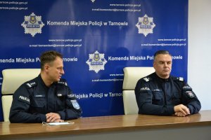 Komendant Wojewódzki Policji w Krakowie nadinsp. Michał Ledzion oraz Komendant Miejski Policji w Tarnowie insp. Mariusz Dymura siedzący za biurkiem w tle tablica z logo i danymi Komendy miejskiej Policji w Tarnowie