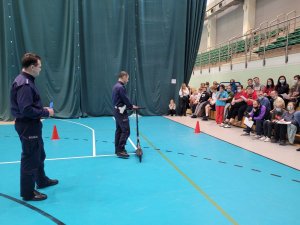 Umundurowani policjanci stoją w hali sportowej w dalszej części zdjęcia uczestnicy siedzący na ruchomej trybunie