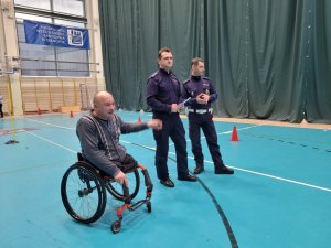 Niepełnosprawny mężczyzna siedzący na wózku inwalidzkim obok stoi dwóch umundurowanych policjantów.