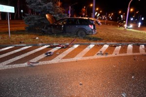 Uszkodzony lewy przedni bok ciemnego samochodu, samochód stoi na trawniku, dookoła porozrzucane plastikowe elementy